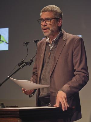Wayde Compton stands at a lectern and addresses the audience while holding a stack of papers in his right hand. Wayde wears glasses, and a brown blazer layered over a fleece zip-up sweater and a patterned collared shirt. 