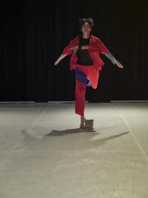 Alexa Mardon leaps towards the camera with their arms outspread in a low V-shape. They wear an unbuttoned red shirt and matching pants with blue detailing. They dance on a white Marley floor.