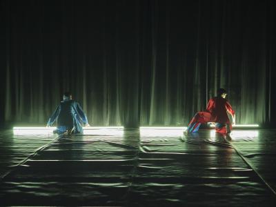 Two people crouch on a Marley dance floor with their backs to the camera. They wear matching blue and red pants and button up tops. They reach towards two fluorescent light tubes that illuminate the space. The image has an otherworldly green colour balance.