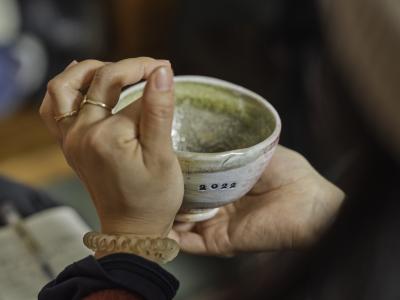 Hands hold a ceramic bowl with 2022 stamped into the side. The person wears gold rings and a clear spiral hair tie around their wrist.