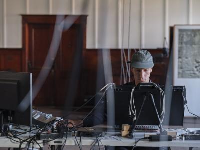 Alexi Baris sits in front of an intellijel synth patch and computer monitor positioned on a grey folding table. He wears a green baseball cap. 