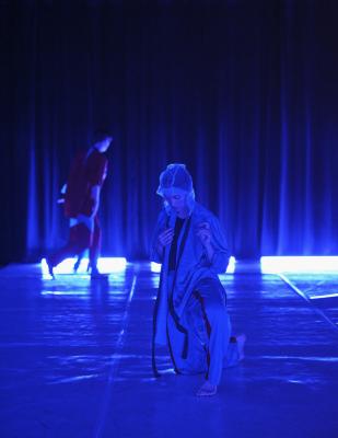 Erika Mitshuhashi kneels and gazes towards the ground. A sheer piece of fabric covers her head. Alexa runs along the back of the dance floor. The whole image is basked in blue light. 