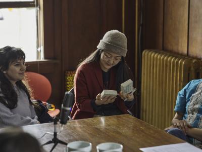 Josephine Lee sits at a wood table and holds the base and lid of a ceramic box in either hand. She wears a taupe toque, glasses, and a red wool coat. Amelia Butcher sits next to her and smiles while gazing across the room. A podcast microphone is positioned on the table between them.