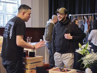 A customer wearing over ear headphones, a green baseball cap, a black Arcteryx jacket, and a backpack, smiles at Derrick Wong, who is shown with his back to the camera. They stand on opposite sides of a table with a display of leather goods.
