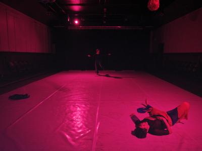 Alexa Mardon lies on a Marley dance floor in the Grand Luxe Hall. In the background, Erika Mitsuhashi dances with her arms outstretched. The room is flooded in fuschia light.