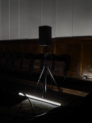 A tubular lighting fixture positioned on the floor illuminates a square speaker on an adjustable stand.
