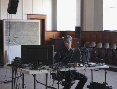 Alexi Baris sits at a grey folding table in front of various electronic music equipment. He wears a baseball cap and long-sleeve button up shirt. Behind him, a speaker is suspended from the ceiling. The windows of the Grand Luxe Hall are open, and bright light floods into the space. 