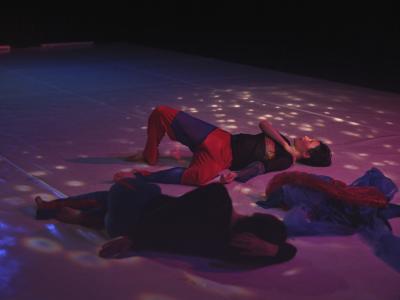 Alexa Mardon and Erika Mitsuhashi lie on a Marley dance floor under soft dappled light. Red and blue tulle garments are piled between them.
