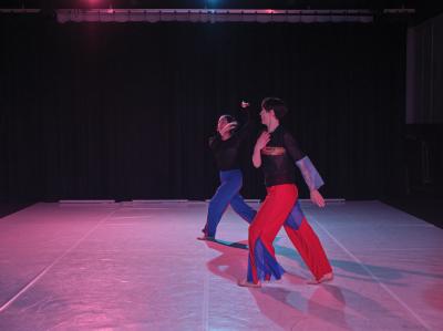 Dancing in sync, Alexa Mardon and Erika Mitsuhashi step forward with their left legs. They are dressed in black tops and red and blue pants with inverse detailing to match. They dance on a Marley floor in the Grand Luxe Hall that is illuminated by pink and blue stage lights.