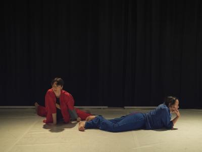 Alexa Mardon and Erika Mitsuhashi lie on their stomachs on a white Marley dance floor. Alexa faces the camera and is dressed in  a matching set of red pants and a button-up top. Erika lies perpendicular to them and is dressed in a similar top and pant combo, but in blue. 