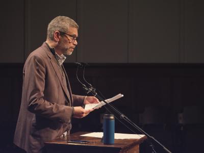 Wayde Compton stands at a lectern, where a blue water bottle, iPhone, and speaking notes are placed. He reads off a piece of paper held in both hands, and gazes towards the audience. Wayde wears glasses and is dressed in a brown blazer