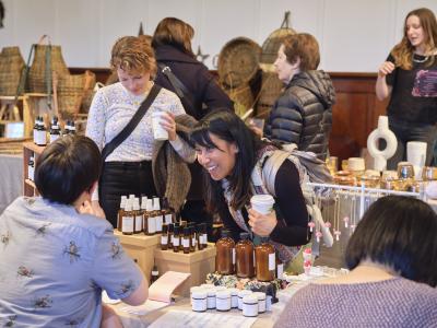 A woman holding a disposable coffee cup smiles and leans toward a vendor seated at their table, which displays a variety of small amber bottles.
