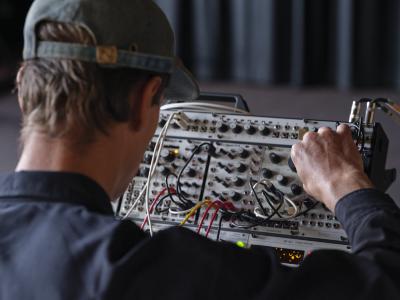 Alexi Baris turns a dial on a synth patch. His back is to the camera, and he wears a baseball cap and a long-sleeve button up shirt. 