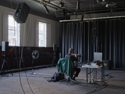 Alexi Baris works on a synth patch set up on a grey folding table, alongside other electronic music equipment. He sits with his back to the camera, and a green jacket is draped on his chair. Four speakers are set-up in the room: two on the floor, and two hanging from the ceiling. 