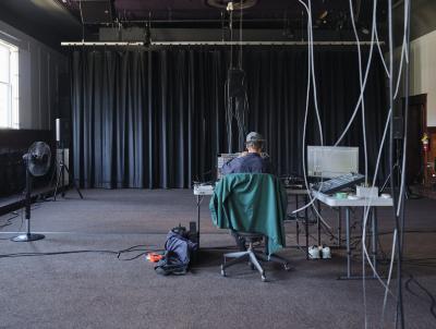 Alexi Baris works on a synth patch set up on a grey folding table, alongside other electronic music equipment. He sits with his back to the camera, and a green jacket is draped on his chair. Cables from a speaker suspended from the ceiling dangle in the camera’s line of sight.