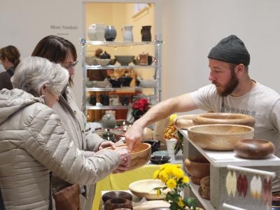 Wearing a grey toque and graphic T-shirt, Tony Dubroy leans over his table display of wooden bowls to show two customers a detail in a piece they are holding.