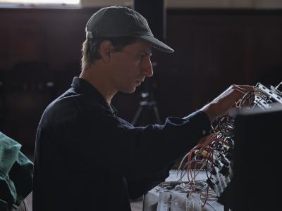 Alexi Baris is seen in profile while working on a synth patch. He is seated, and wears a long-sleeve button up shirt and a baseball cap.