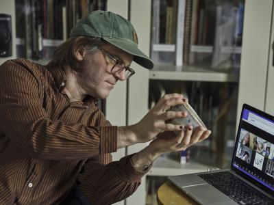 Christian Newby holds a ceramic bowl up to a laptop that is open on a Zoom call. Christian wears glasses, a green ball cap and a brown striped shirt. His nails are painted various shades of red.