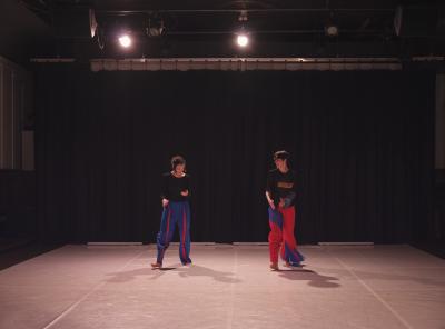Erika Mitsuhashi and Alexa Mardon stand side-by-side on a Marley dance floor, smiling while engaging in a duet. They are dressed in black tops and red and blue pants with inverse detailing to match. 