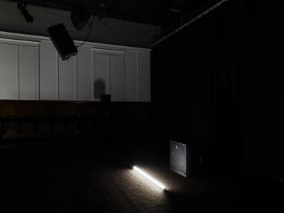 A tubular lighting fixture positioned on the floor illuminates a large box-like speaker sitting next to it. Another speaker is suspended from the ceiling.