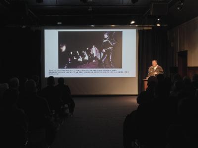 Michael Dang stands at a lectern on the stage of the Grand Luxe Hall and is illuminated by a spotlight. An archival photo is projected on the screen next to him that shows John Dowd in a leather jacket and pants, a black t-shirt that said FETISH, and a shark fin bathing cap. In the background, Pascal performs in a pink robe. She is surrounded by several male dancers, also wearing shark fin hats. The accompanying caption reads: Pascal, John Dowd etc., performing at the Decca Dance, 1974, Morris/Trasov archive at the Morris & Helen Belkin Art Gallery, followed by the image accession number. 