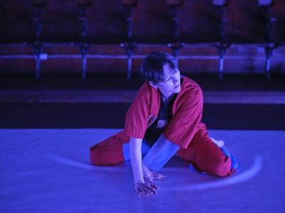 Alexa Mardon kneels on a Marley dance floor with their hands twisted in an almost crawling motion. They wear a red button up shirt and pants with blue detailing.