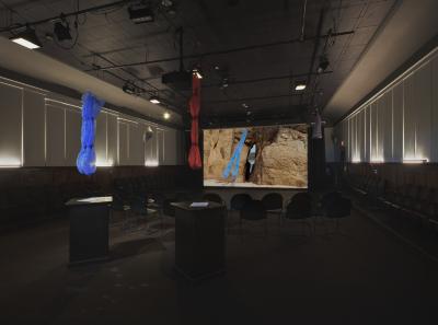 Two tulle sleeves, one red and one blue, hang from the ceiling of the Grand Luxe Hall. These garments are echoed in an image projected on a screen at the back of the space that shows a blue tulle garment dangling in front of a rock face. A number of chairs are set up facing the screen. 
