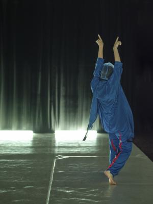 With her back to the camera, Erika Mitsuhashi points towards the ceiling. She wears a satiny blue robe and matching pants with a red stripe down the back of the legs. Her head is covered by a sheer piece of blue fabric.