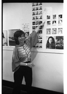 A person wearing a dolman-sleeve shirt addresses their attention to the camera, and smiles while pointing with their left hand to a polaroid image on the wall. In their right hand they hold a glass goblet.  
