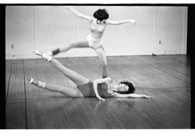 Two women perform a dance in a studio. They both have short hair, wear leotards, and leg warmers. One dancer lays on her side with one arm stretched out as the other bends to touch the floor and her legs fan out in the air. The other dancer stretches her arms and leaps in the air.