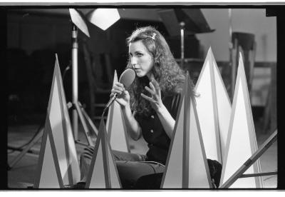 A woman sits criss-cross as she holds a microphone in one hand as she gestures with the other as she performs. She is encompassed by several cone-like props of different heights. Behind her are other pieces of equipment; a music stand, stage light, and chairs.