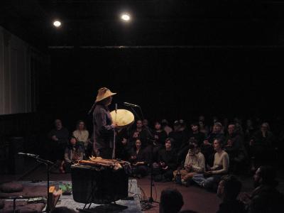 Seen in profile, Paul Chiyokten Wagner stands while playing a Coast Salish drum. Paul stands next to a table with a variety of hand carved woodwind instruments, and he is dressed in a woven cedar hat. The stage area is demarcated by a blue carpet, and the audience is seated on the floor and in chairs arranged in a curve.  