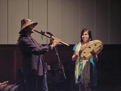 Paul Chiyokten Wagner plays a wooden flute. He wears a woven cedar hat adorned with a woven cedar rose, abalone shell, and feather. The hat casts his face in dappled shadows. He also wears a blue tunic and beaded necklace. 
Jennifer Ambriz gazes towards him while playing a Coast Salish drum. She wears a blue dress with yellow flowers, and a white tassel scarf.  