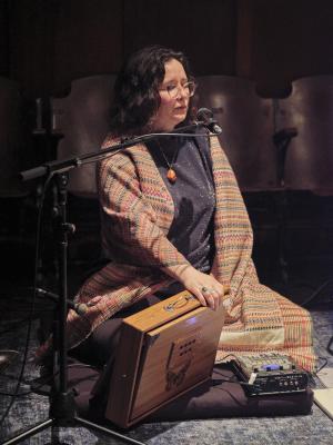 Jessika Kenney sits crossed legged on a meditation pillow on a blue carpet. She sings into a microphone while playing a shruti box with her right hand. She wears glasses, a geometric pendant, and a sparkly top under a woven shawl. 
