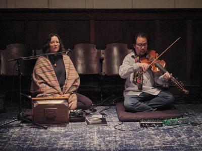 Jessika Kenney and Eyvind Kang sit side by side on meditation pillows on a blue carpet. Jessika’s eyes are closed as she vocalizes into a mic. She wears glasses, a geometric pendant, and a sparkly top under a woven shawl. 
A shruti box, sample pad, and several books are arranged in front of her. Kang plays viola d’amore and wears glasses, a floral scarf, blue button-up shirt, and blue jeans. 