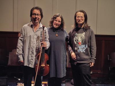 A group photo of musicians Eyvind Kang and Jessika Kenney with curator Aki Onda. Eyvind wears glasses and a blue button-up shirt, and holds his viola d’amore. Jessika, with her arms around Eyvind and Aki, wears glasses, a blue sparkly dress, and a geometric pendant. Aki also wears glasses, as well as a long sleeve sweater printed with a graphic of a tiger.
