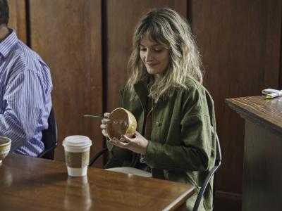 Lauren Lavery sits at a wood table and holds a small yellow ceramic bowl, and a green pencil. She wears a green chore coat and her nails are painted blue.