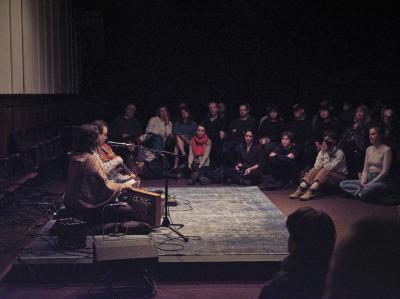 Jessika Kenney and Eyvind Kang sit next to each other on a raised platform covered in a blue carpet. A microphone, various cables and effects pedals, and a shruti box are arranged around them. Jessika vocalizes as Eyvind plays viola d’amore. The musicians are surrounded by audience members who are seated on the floor and in chairs. The Grand Luxe Hall is very full. 