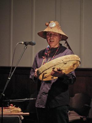 Paul Chiyokten Wagner sings while playing a Coast Salish drum. He wears a woven cedar hat adorned with a woven cedar rose, abalone shell, and feather. The hat casts his face in dappled shadows. He also wears a blue tunic and beaded necklace. 