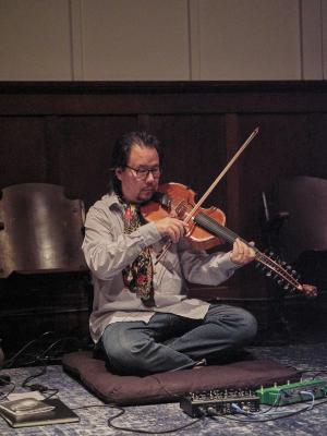 Eyvind Kang sits crossed legged on a meditation pillow on a blue carpet while playing viola d’amore. He wears glasses, a floral scarf, blue button-up shirt, and blue jeans. 