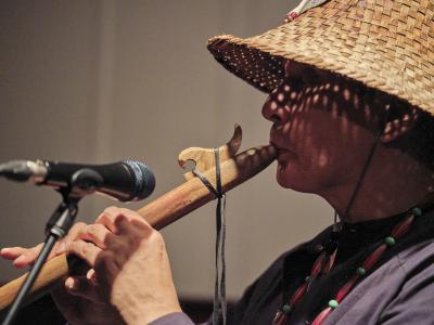 Paul Chiyokten Wagner is seen in profile while playing a wooden hand carved flute. A microphone is pointed towards the instrument. Paul wears a woven cedar hat that casts his face in dappled shadows.