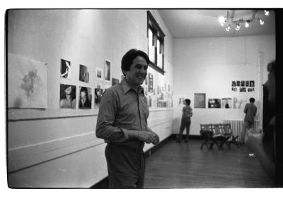 A person in the foreground smiling in front of a wall of portraits. In the background, two people address their attention to the exhibited images.
