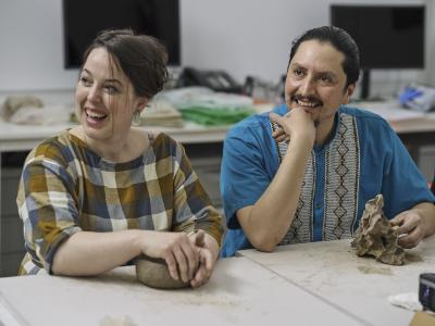 Two guests smile at something outside of the frame. They keep their hands working on the slabs of clay in front of them.
