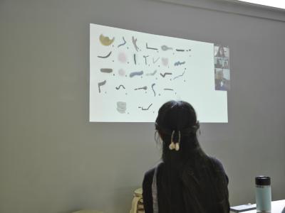 One guest faces a projection of a Zoom meeting. The presentation shows paint markings in different shapes and colours. Each shape is labeled by a different letter A through Z.
