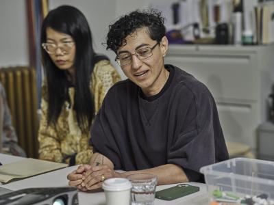 An artist in a black sweater and reading glasses play with their fingernails while speaking. They tilt their head to the right while looking to the left. Another guest in a beige top sits next to them, listening.
