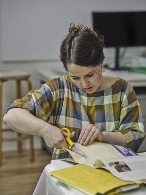 A guest wearing a plaid top cuts indiscernible lines of text from a magazine. They pull the page taut with their right hand while cutting forward with their left. 
