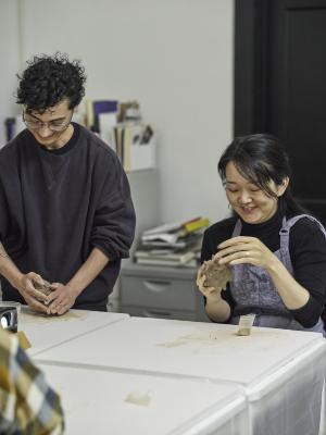 Two guests stand on one side of a table shaping clay. An artist in a black sweater pushes the clay on the table, while the other, in a blue apron, pulls the clay with their fingers.