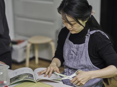 A guest wearing an apron cuts a strip of indiscernible words from a magazine. They carefully rest their left hand on the page while lining up the blades of soft grip scissors to create their slice.
