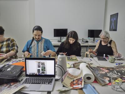 Guests sit around a table cluttered with electronics, magazines, paper towels, glue sticks and scissors. They work away, each artist concentrating on the material in front of them. An open laptop reveals a zoom meeting with multiple participants.