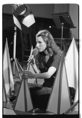 A woman sits criss-cross as she holds a microphone while performing. She is encompassed by several cone-like props of different heights and holds on to one of them. Behind her are other pieces of equipment; a music stand, stage light, and chairs.